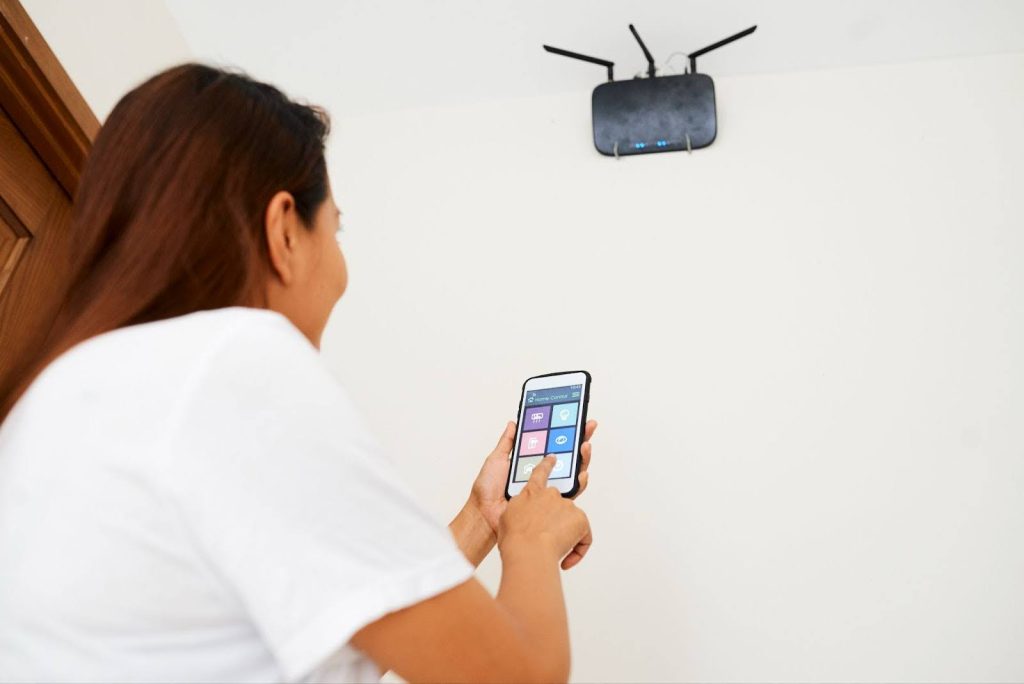 A woman uses a smartphone app to control a black Wi-Fi router mounted high on a white wall