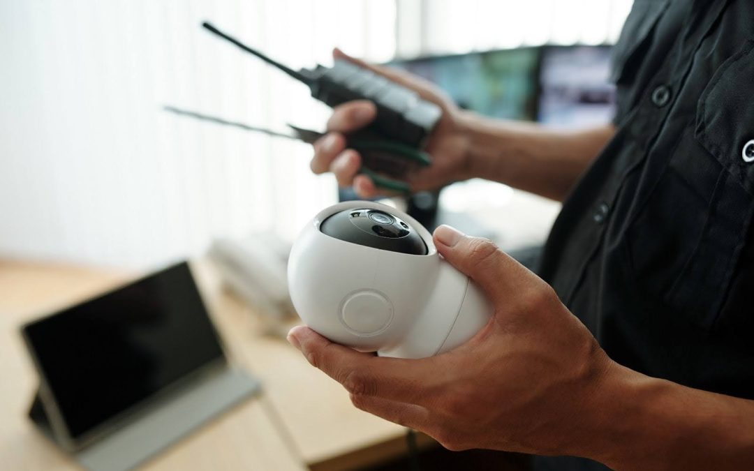 A close-up of a person's hands, likely a security professional, holding a white CCTV security camera and a walkie-talkie