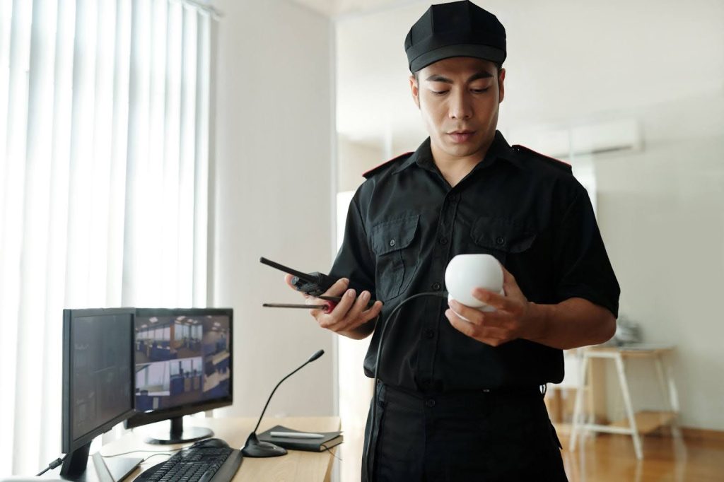 A security guard in a black uniform and cap holds a white security device and a walkie-talkie, standing near a desk with monitors displaying CCTV footage.